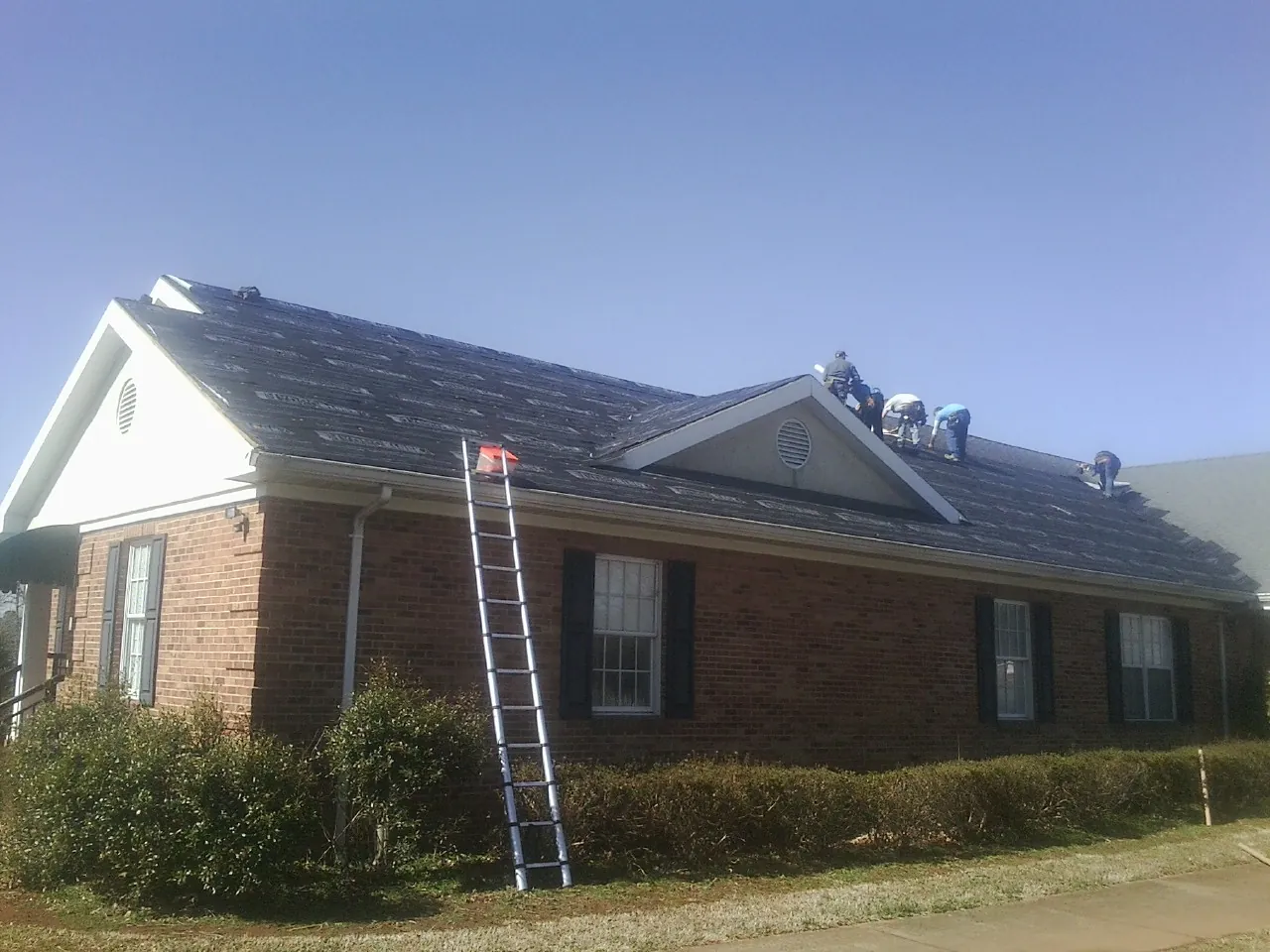 Crew tearing shingles off a brick ranch roof with a ladder set — Emergency Roof Repair work by our crew in Cofield