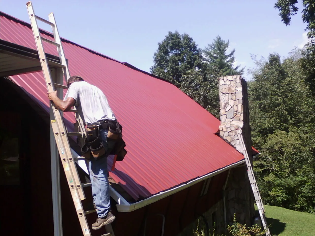 Roofer on a ladder beside a red metal roof and stone chimney — Soffit & Fascia Repair by Roofing Brothers in Cofield